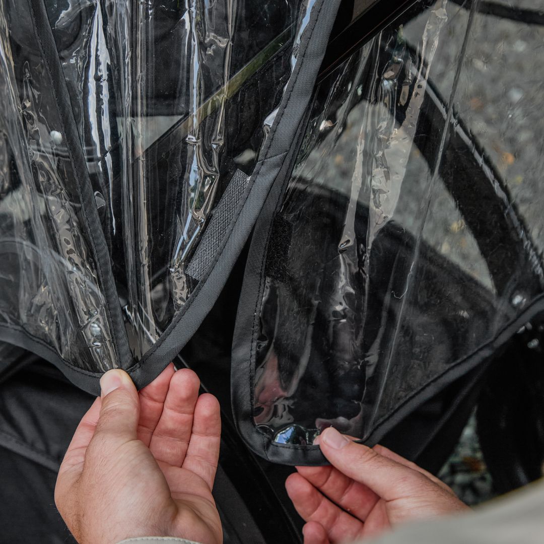 Close-up of hands adjusting the Pram Rain Cover on a stroller with a black frame, an outdoor background visible through the transparent waterproof material.
