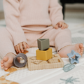 A baby in a pink outfit sits on a play mat, reaching with bare hands and feet for colorful pieces of the Nuby Shape Sorter Learner Toy, building hand-eye coordination while exploring stacking activities.