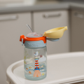 A Nuby UK Super Straw Twin Pack kids’ water bottle with a straw, seagull and lighthouse design, and light blue and orange lid with open yellow cover sits on a white kitchen surface.