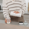 A person in a striped sweater pours liquid from a white thermos into a baby bottle on the kitchen counter, with the Nuby UK RapidCool Bottle Maker and Portable UV Steriliser Bundle, plus bottle cap and nipple, beside them.