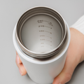 A hand holds a white metal cup from above, showing silver measurement markings (60–240ml) inside—ideal for use with the Nuby UK RapidCool Bottle Maker and Portable UV Steriliser Bundle. The inside rim and measurements are silver.