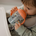 A young child in a high chair drinks from a Nuby UK Grip n Sip Cup (Whales & Bees, 4 Pack), a transparent no-spill cup with orange handles and lid, featuring animal and plant designs. The child wears a green outfit with white patterns.