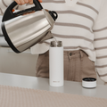 A person in a striped sweater pours hot water from an electric kettle into a white insulated bottle, likely prepped with the Nuby UK RapidCool Bottle Maker and Portable UV Steriliser Bundle, on a kitchen counter, the cap set nearby.