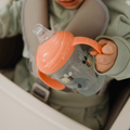 A baby in a high chair, dressed in light green and secured by a harness, holds a Nuby UK Grip n Sip Cup from the Whales & Bees 4 Pack, featuring orange handles and bee and flower designs—one of the best no-spill sippy cups.