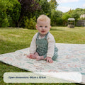 A baby sits on the Fold N Go Play Mat—a patterned cushioned foam mat—outdoors on grass, with trees and a well in the background. Text on the image reads: Open dimensions: 90cm x 121cm.