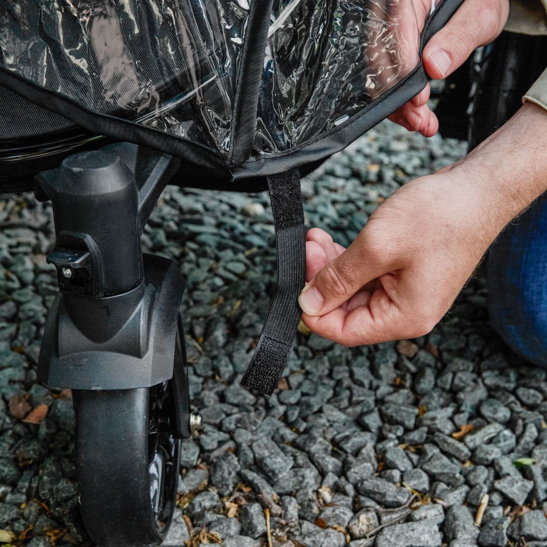 A person fastens a black strap to secure the Pram Rain Cover onto a stroller wheel while outdoors on a gravel surface.