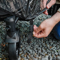 A person fastens a black strap to secure the Pram Rain Cover onto a stroller wheel while outdoors on a gravel surface.