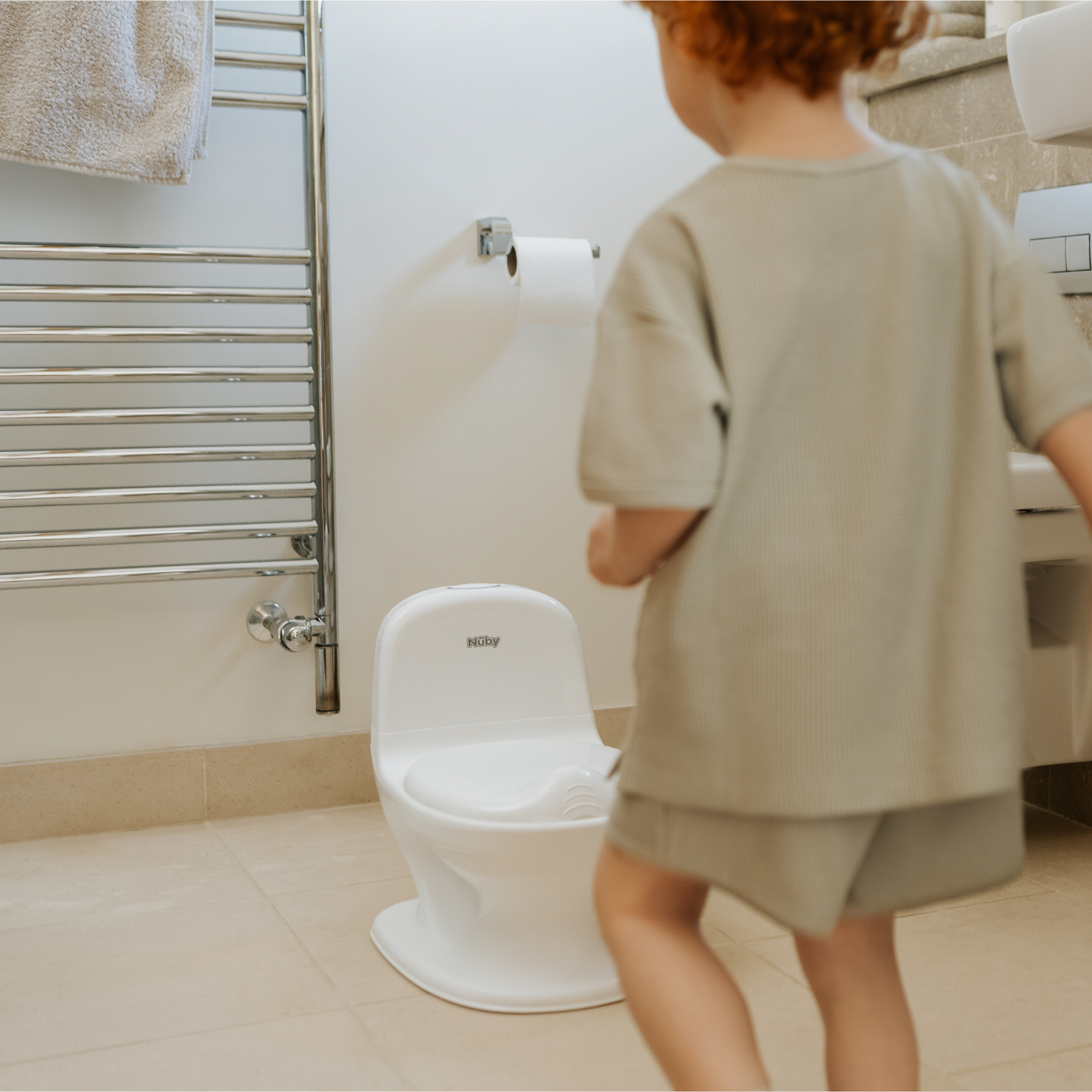 A young child with curly hair stands in a bathroom near the Nuby UK Potty Training Kit Bundle mini toilet, facing away from the camera, with a towel rack, toilet paper holder, and beige tiles in view.