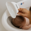 An adult pours water over a baby’s head with a white Nuby cup while the baby sits in the Nuby Sit Me Up Baby Bath, making bathtime safe and convenient. The baby’s back and head are visible, and the adult’s hand grips the cup.