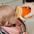 A baby in a pink bib drinks from the Nuby All Around Cup 2 Pack - Pink, a spill-proof cup with yellow handles, orange body, and 360-degree valve, as an adult helps hold the two-handled cup.