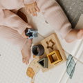 A baby in pink ribbed clothes plays on a patterned mat, holding a blue star-shaped block. Nearby, the Nuby Shape Sorter Learner Toy with star, circle, and triangle pieces helps build hand-eye coordination.