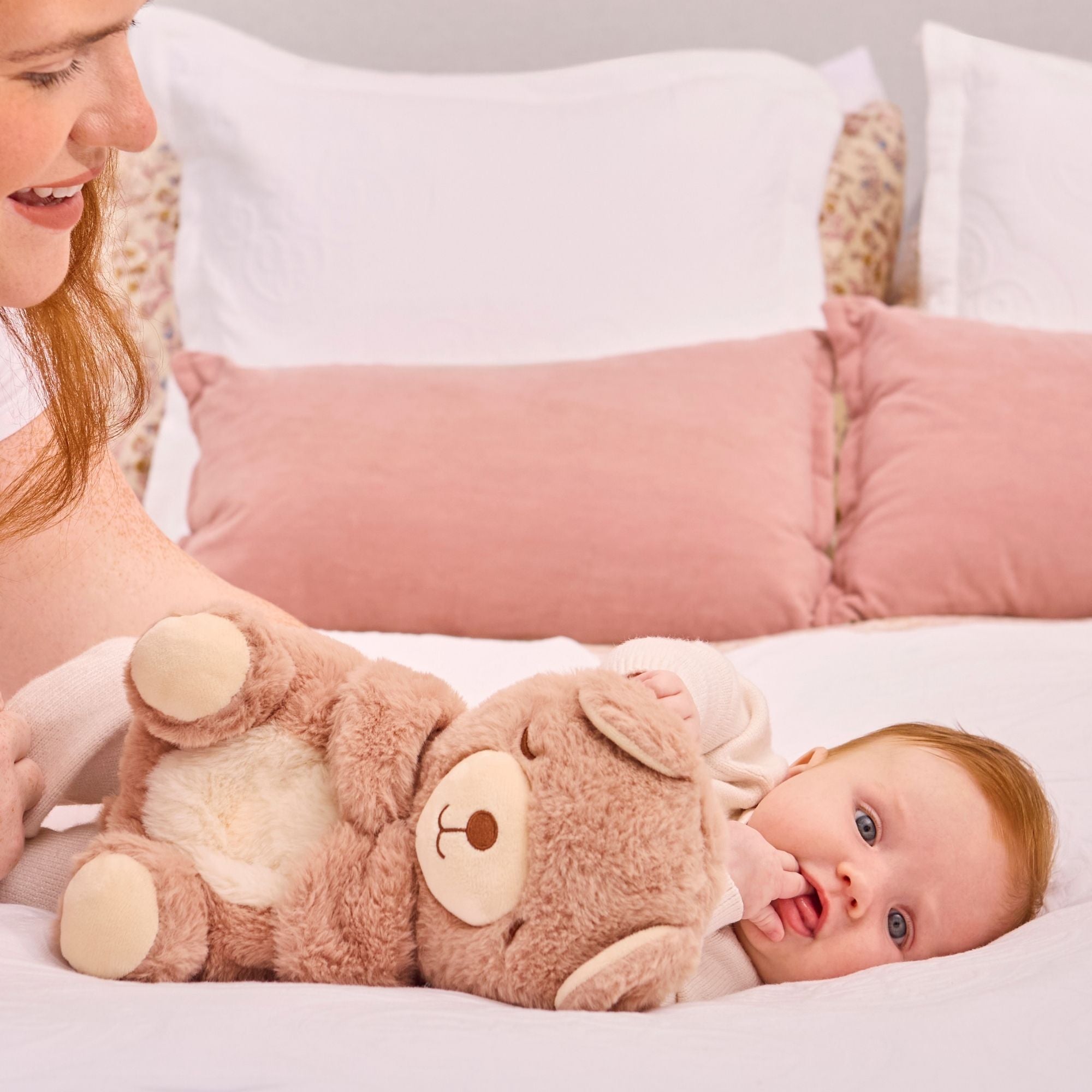 A baby with light brown hair lies on a bed with white sheets and pink cushions, holding the Biscuit Bear Calming Comforter plush while a woman sits beside them.