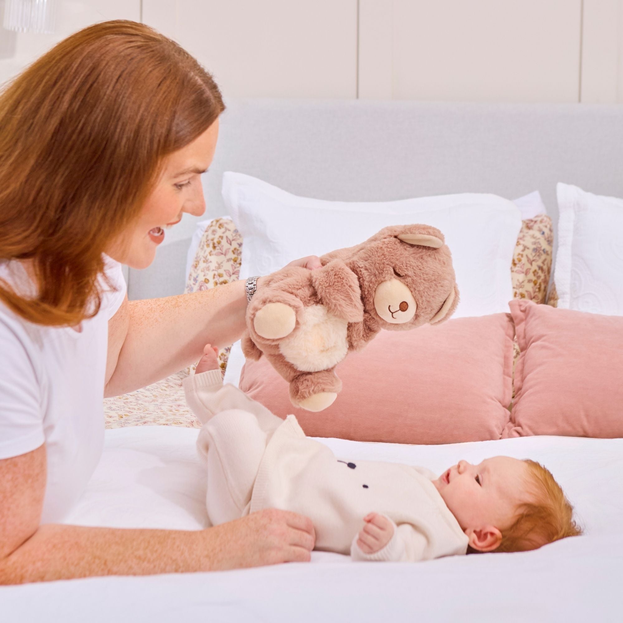 A red-haired woman smiles as she holds the Biscuit Bear Calming Comforter above a baby lying on a bed, both surrounded by pillows and white bedding.