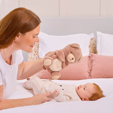 A woman smiles as she holds the Biscuit Bear Calming Comforter plush above a baby in a light outfit, who gazes up at the toy amid white and pink pillows on the bed.