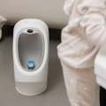 A small white Nuby My Real Urinal with a blue spinning target is set on a gray floor beside light furniture and a beige blanket—ideal for urinal and potty training.
