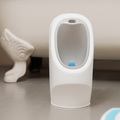 The Nuby My Real Urinal, a white plastic potty training urinal with a blue spinner, is perfect for urinal training and stands on a light bathroom floor near a clawfoot tub.