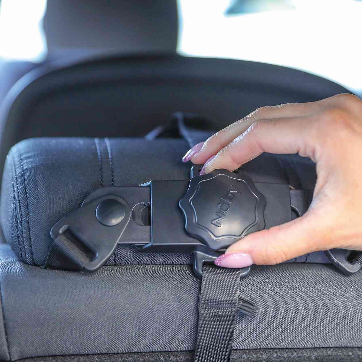 A hand adjusts a headrest strap with a knob labeled IGNY, securing the shatter-resistant Safe and Secure Baby Car Mirror to the back of a car seat.