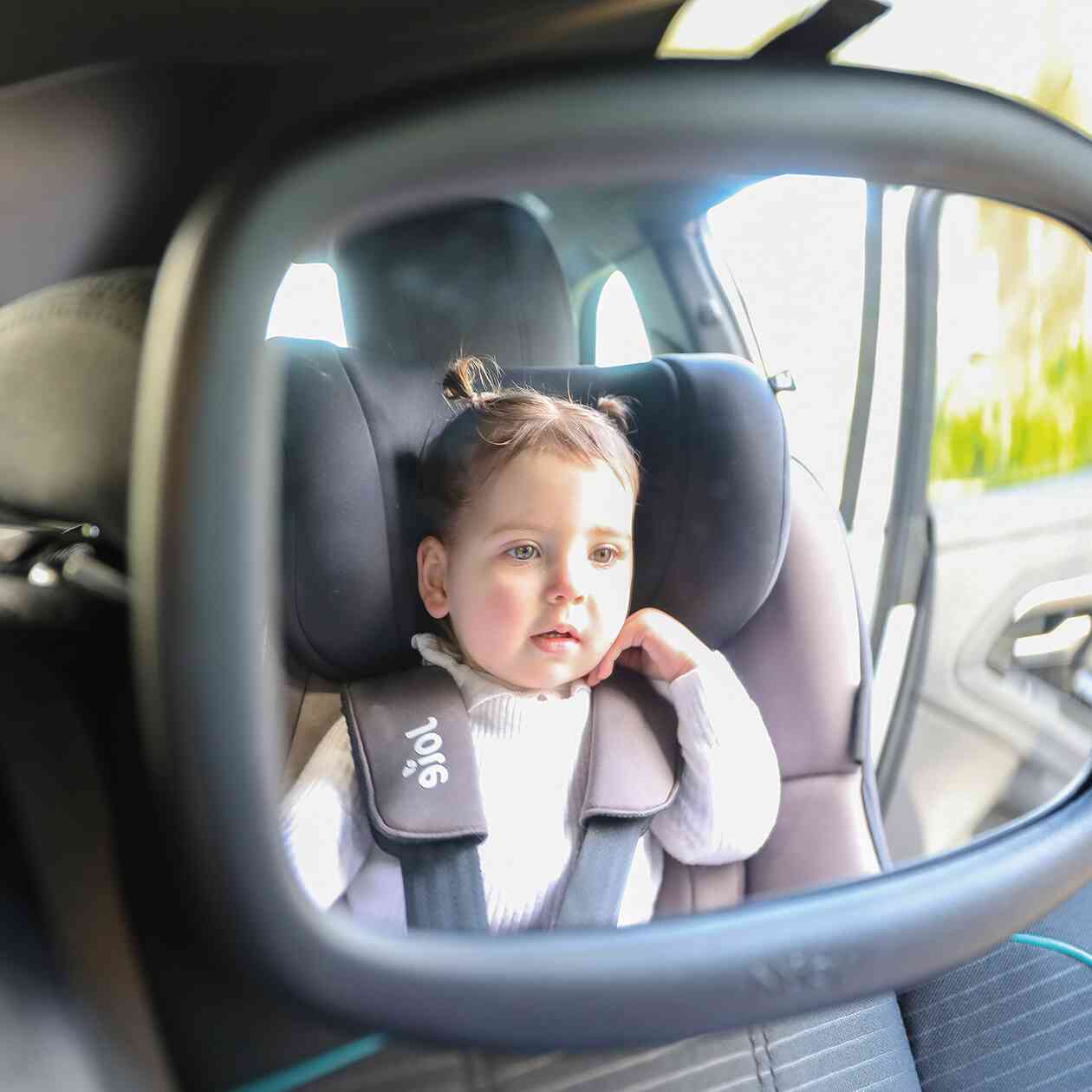 A toddler in a white sweater and pigtails sits rear-facing, visible through the Safe and Secure Baby Car Mirror's shatter-resistant glass, gazing out curiously.