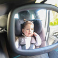A toddler in a white sweater and pigtails sits rear-facing, visible through the Safe and Secure Baby Car Mirror's shatter-resistant glass, gazing out curiously.