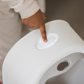A child’s hand presses the white, oval Next button on the Nuby My Real Urinal, a potty training device with a smooth plastic surface and a circular hole below the button.