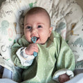A smiling baby in a green bib sits in a high chair, holding a blue Nuby Dinky Dippers Baby's First Spoon. The high chair cushion displays animal drawings and words like "Hippo" and "Cat" on a light backdrop.