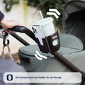 A hand pushes a stroller with the Nuby Pram Cup & Phone Holder attached to the handle, securing both a cup and phone. Text at the bottom: 2-in-1 cup and phone holder—essential on-the-go pram accessory by Nuby.