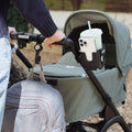 A person pushes a green stroller outdoors on a gravel path, featuring the Nuby Pram Cup & Phone Holder attached to the handle, with a gray backpack on the stroller—highlighting useful pram accessories.
