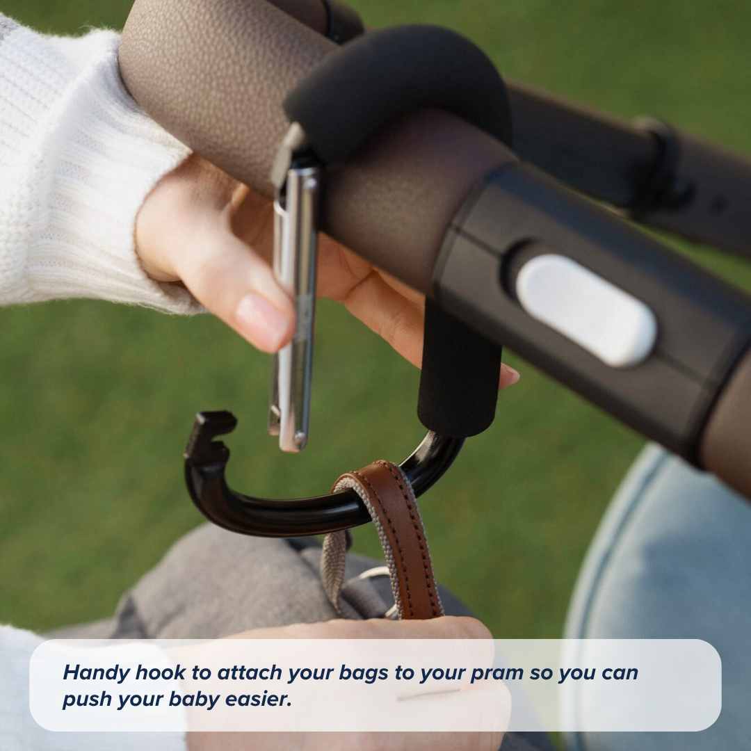A person uses the Nuby Handy Pram Hook to attach a brown leather bag to a stroller handle outdoors on grass. Text reads: Make outings easier—secure your bags to your pram with the Nuby Handy Pram Hook!.