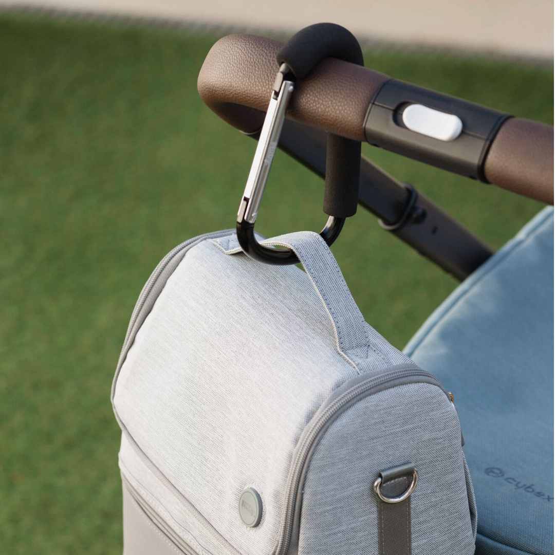 A close-up shows a Nuby Handy Pram Hook in black holding a light gray bag on a stroller’s brown handle, with green grass in the background—highlighting this practical accessory for parents.