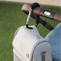 A close-up shows a Nuby Handy Pram Hook in black holding a light gray bag on a stroller’s brown handle, with green grass in the background—highlighting this practical accessory for parents.