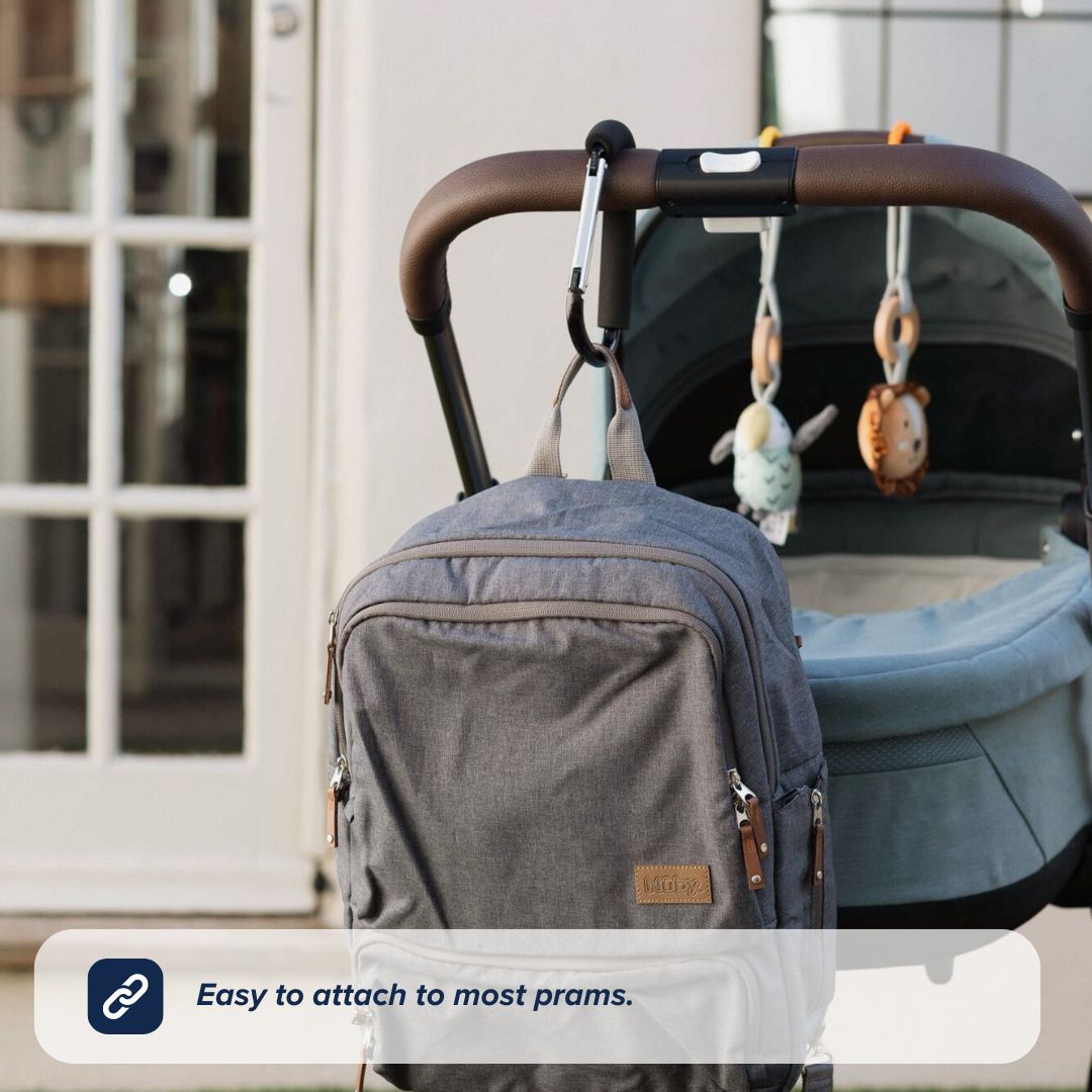 The Nuby UK Pushchair Essentials Kit hangs from a pram hook on the stroller handle, with soft animal-shaped toys above the bassinet. A glass door and white building are in the background. Text: Easy to attach to most prams.