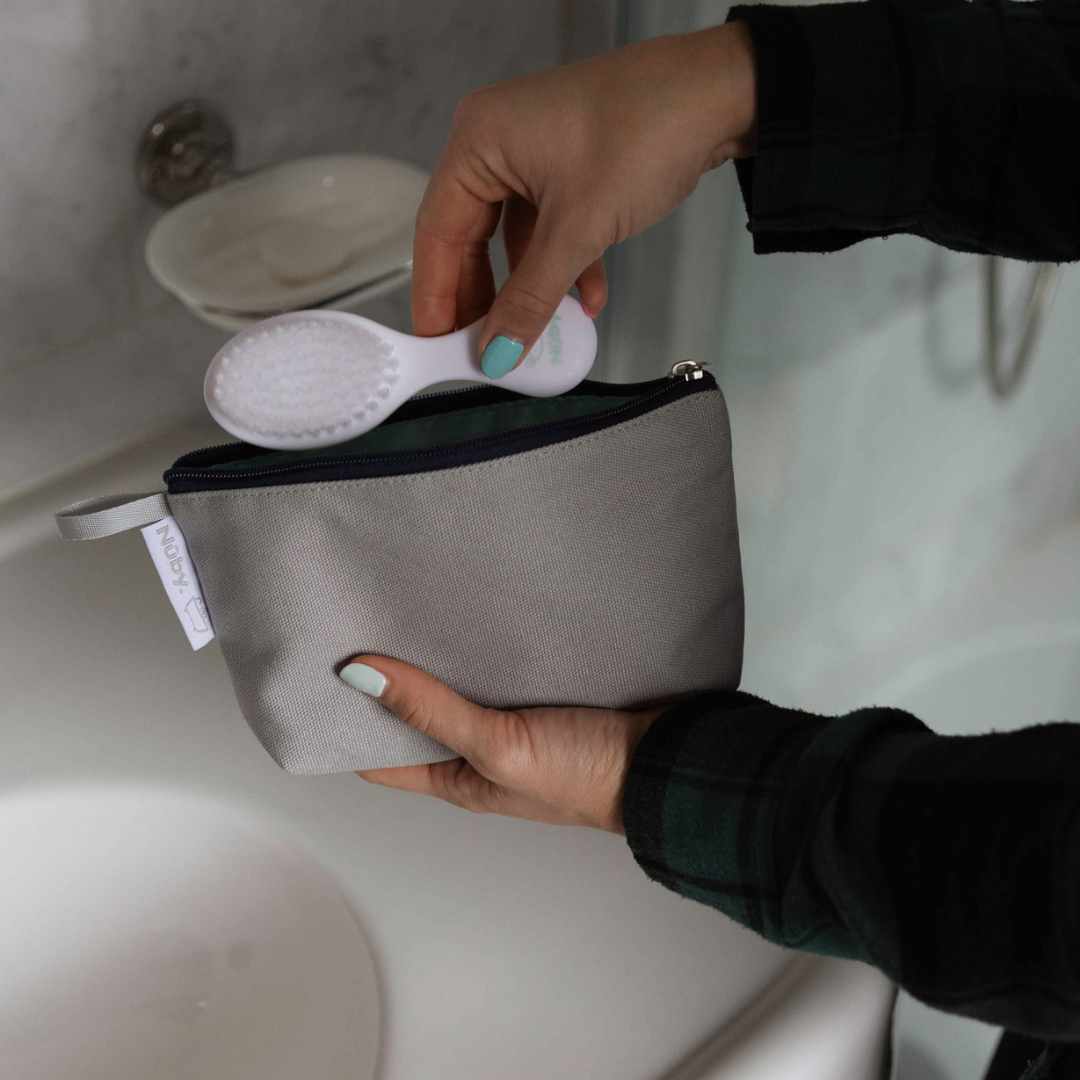 A person places a white facial brush from the Nuby Baby Healthcare and Grooming Kit into a light gray zippered pouch in the bathroom by the sink and soap dish.