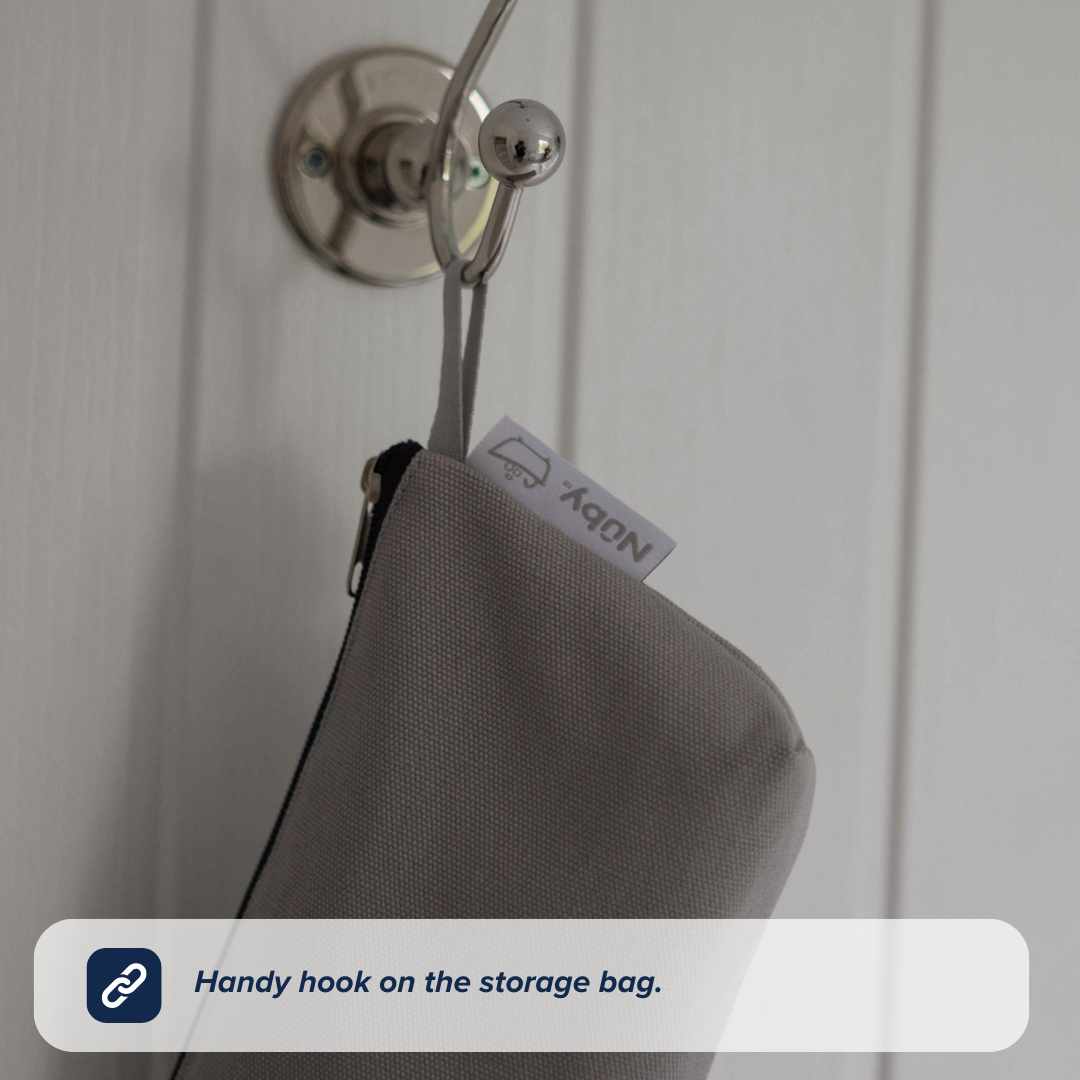 A gray Nuby Baby Healthcare and Grooming Kit bag with a zipper hangs from a metal wall hook. The bag features a white tag with text and a small car icon, making it ideal for storing baby grooming or bath essentials.
