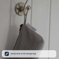 A gray Nuby Baby Healthcare and Grooming Kit bag with a zipper hangs from a metal wall hook. The bag features a white tag with text and a small car icon, making it ideal for storing baby grooming or bath essentials.