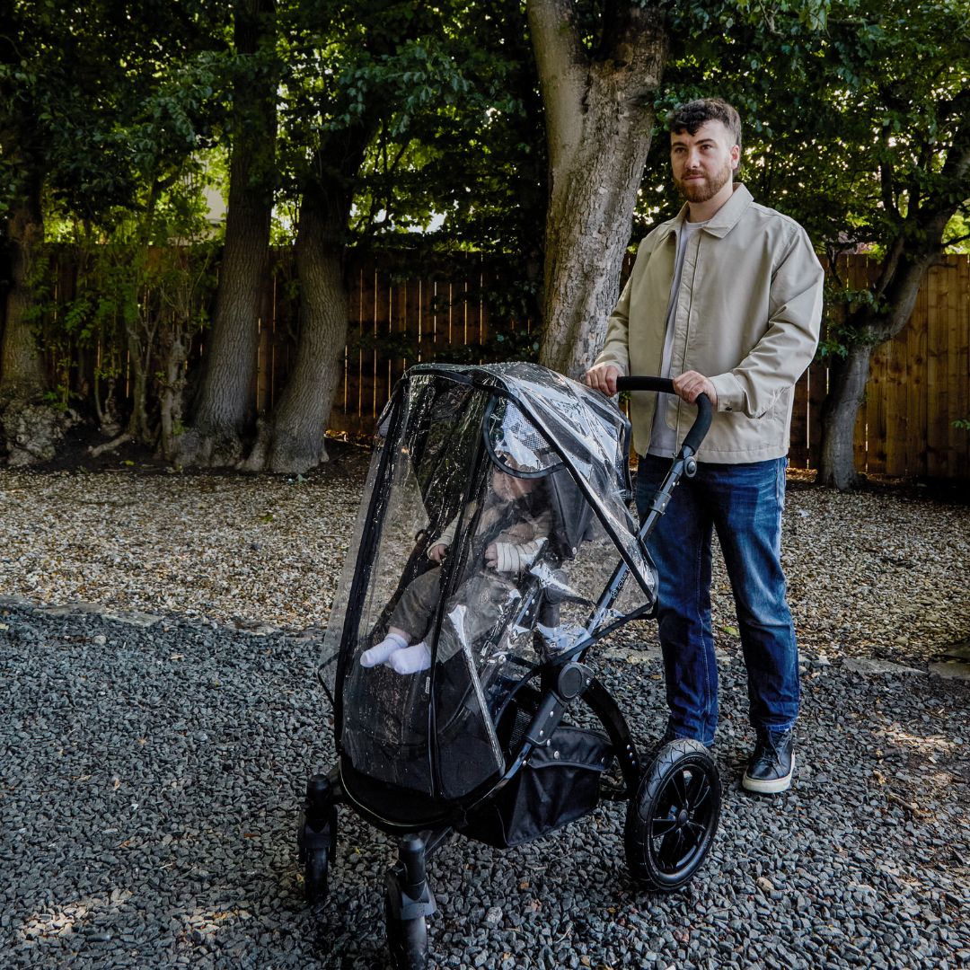A man stands outdoors on gravel, holding a stroller with a seated child, which is protected by the Pram Rain Cover; trees and a wooden fence are visible in the background.