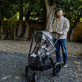 A man stands outdoors on gravel, holding a stroller with a seated child, which is protected by the Pram Rain Cover; trees and a wooden fence are visible in the background.