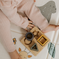 A toddler in pink ribbed clothing sits on an animal-print play mat, playing with the Nuby Shape Sorter Learner Toy, which helps develop hand-eye coordination with circular, star, triangle, and square shapes.
