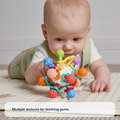 A baby lies on their stomach on a play mat, reaching for the Twist Ball Teether. Text reads: Multiple textures for teething gums.