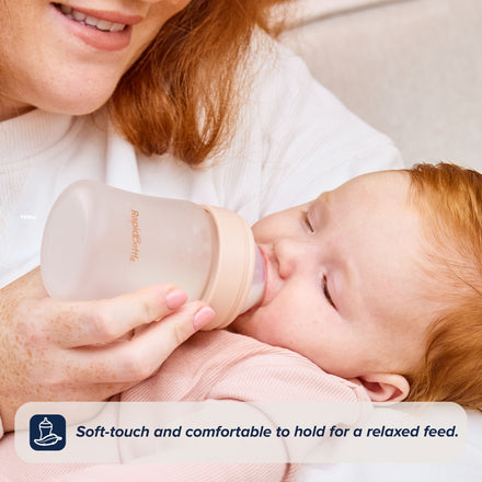 A smiling woman feeds a content baby dressed in pink using the RapidBottle Anti-Colic Glass Feeding Bottle. Text: Soft-touch and comfortable to hold for a relaxed feed.