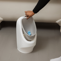 A hand lifts the gray removable inner part of the Nuby My Real Urinal, a white plastic kids potty training urinal with a blue clip, making potty or urinal training easier for children on a light-colored floor.