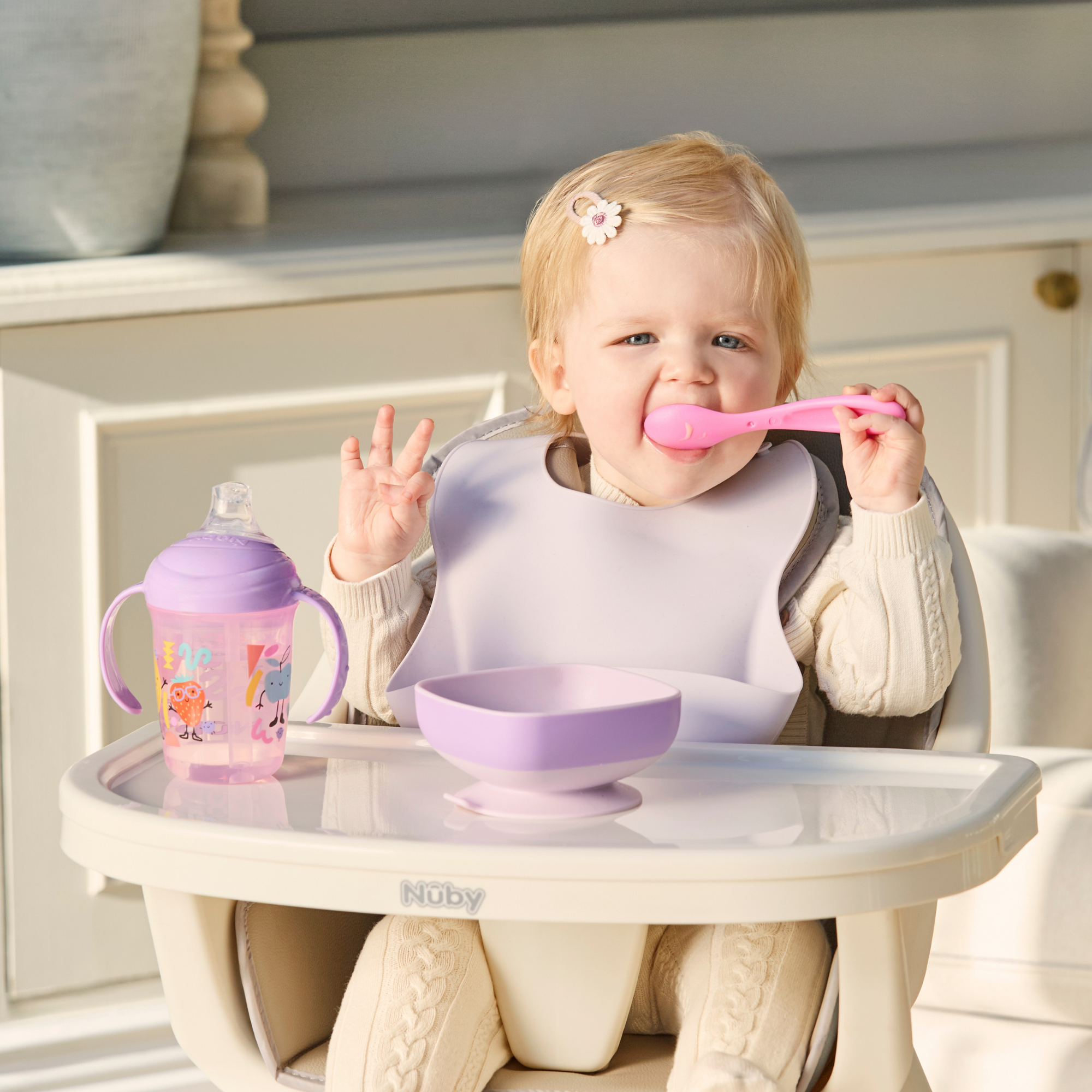 A blonde baby sits in a high chair, wearing a Silicone Bibs 3 Pack bib and holding a pink spoon. On the tray are a purple bowl and a pink sippy cup.