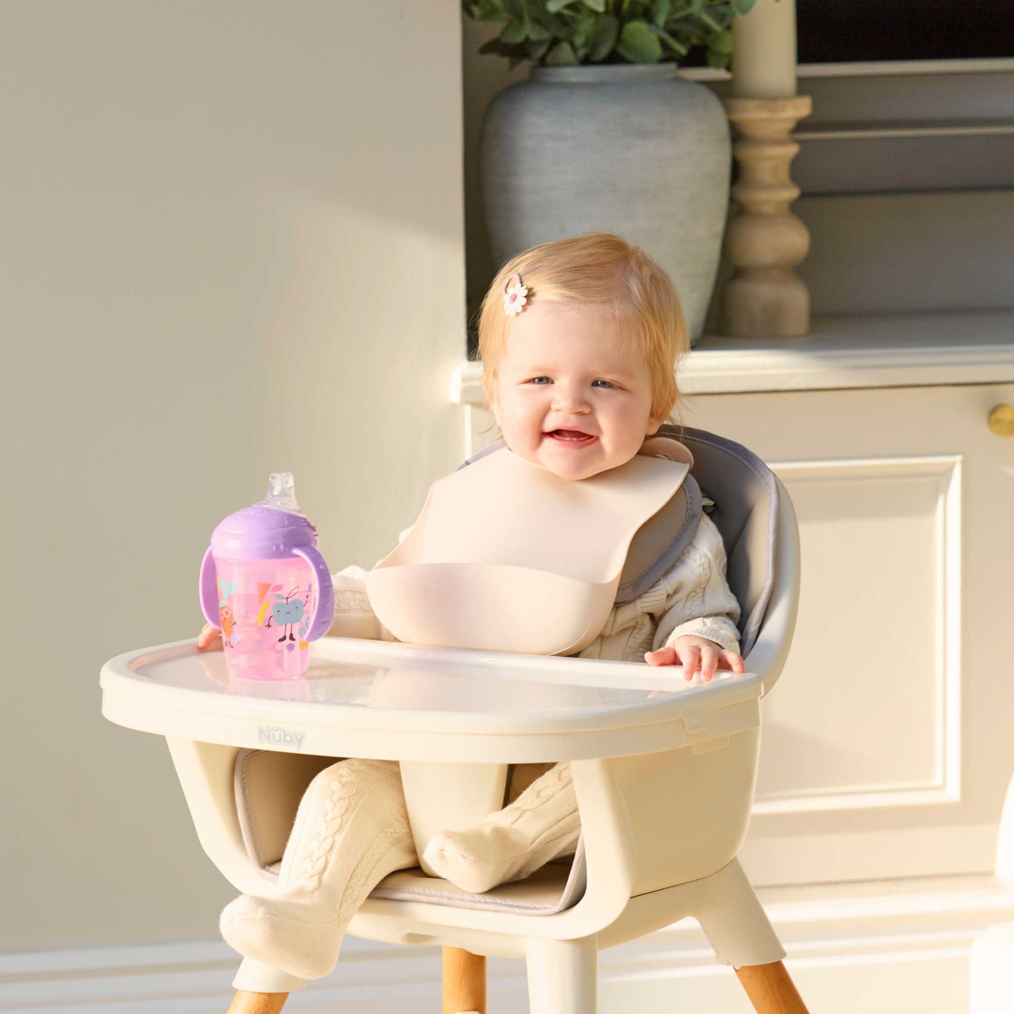A smiling, blonde baby sits in a highchair wearing one of the Silicone Bibs 3 Pack. The baby holds a purple sippy cup and wears a light-colored outfit, with a plant and cabinets visible in the background.
