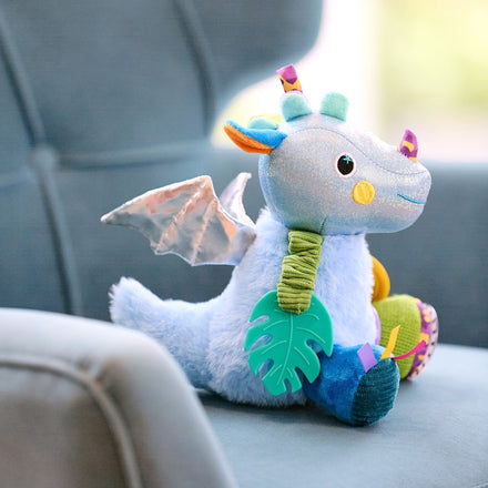 The Things with Wings George Pram Toy is a sensory plush dragon with blue fur, shiny wings, colorful horns, and playful patterned feet. It holds a green leaf-shaped teether and sits on a soft blue armchair—perfect for babies!.