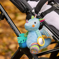 The Things with Wings George Pram Toy, a colorful sensory plush dragon featuring blue fur, green ears, purple horns, and shiny wings with sensory elements, is attached to a black stroller. Autumn leaves cover the ground in the background.