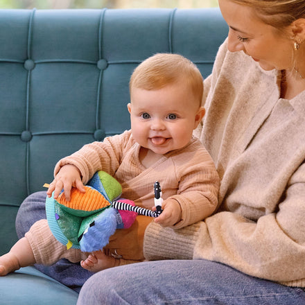 A smiling baby in light brown clothes sits on an adult's lap, holding the Things with Wings Sensory Grab Ball, while the adult smiles down at the baby. They are on a teal sofa in a bright, cozy room.