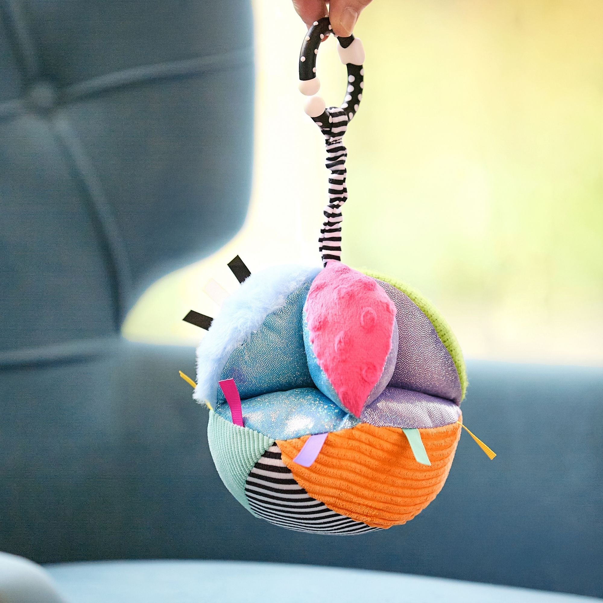 A hand holds the Things with Wings Sensory Grab Ball, a vibrant baby rattle toy with assorted fabrics, patterns, tags, and a black-and-white striped loop. A blue armchair appears blurred in the background.