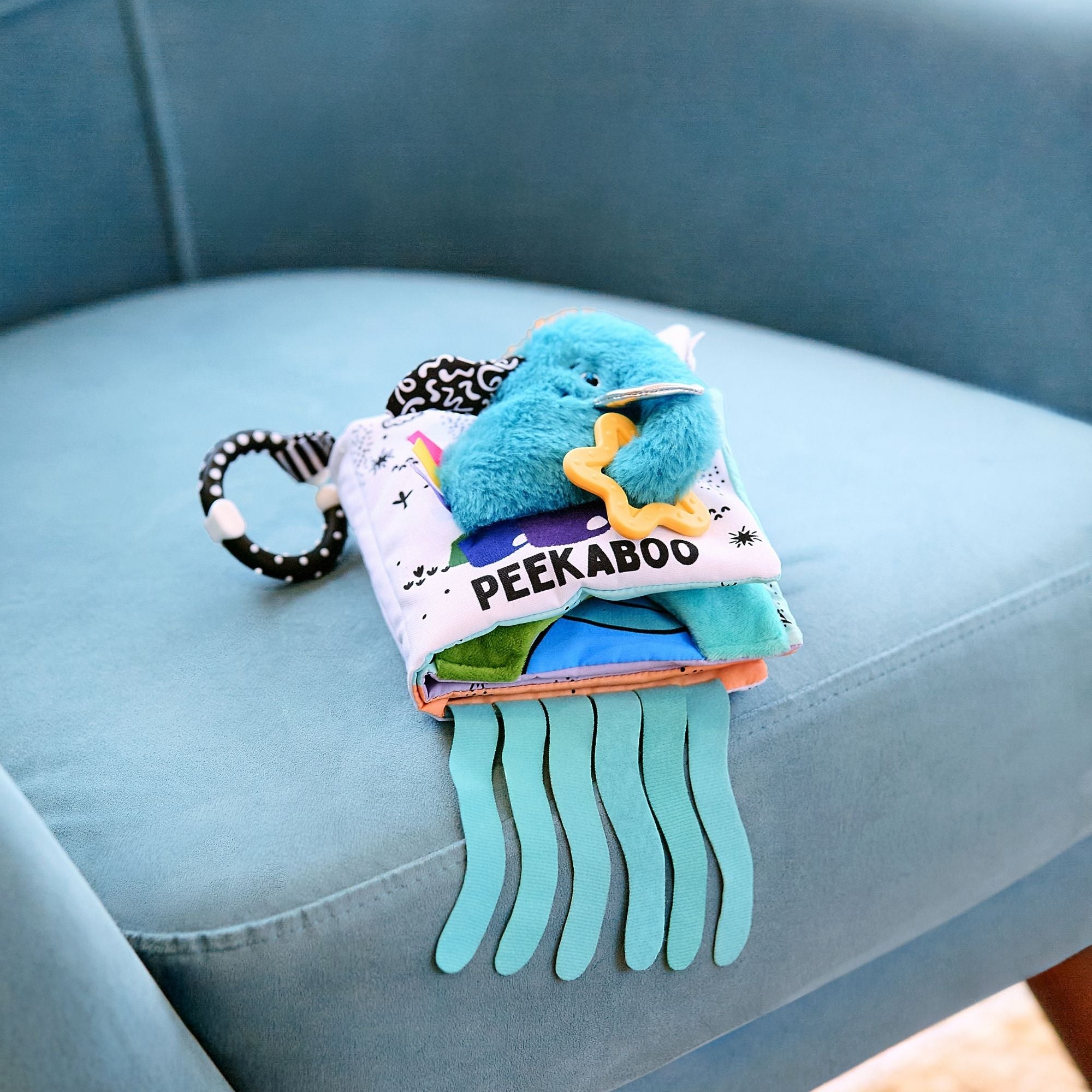 The Things with Wings Peek a Boo Book, a soft baby toy with colorful fabric pages and attached sensory items, rests on a light blue upholstered chair. Blue ribbon-like strips hang from the bottom of the book.