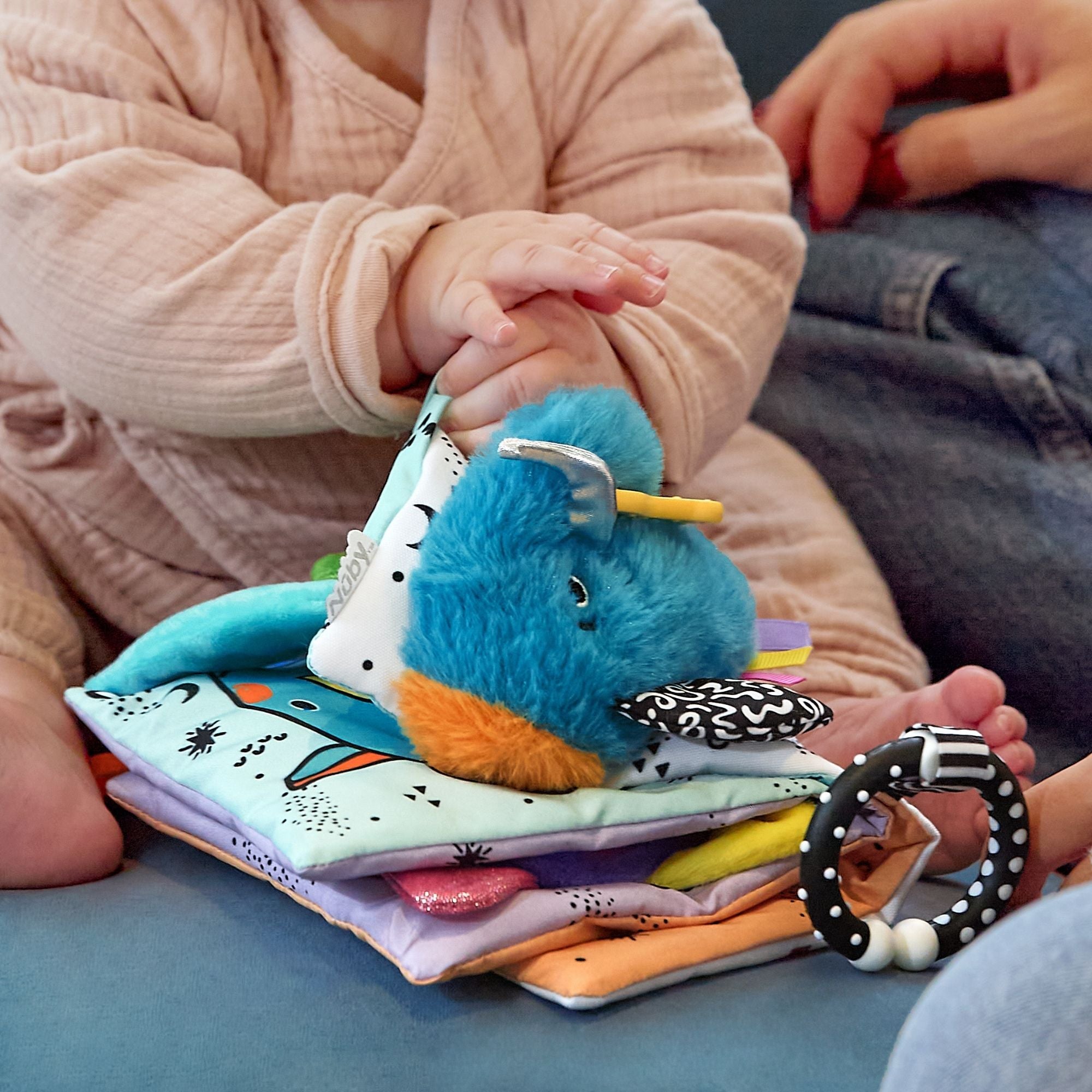 A baby in soft clothes touches the Things with Wings Peek a Boo Book, featuring colorful fabric pages and plush toys, while an adult sits nearby.