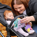 A smiling woman shows the "Things with Wings Peek a Boo Book" to an infant sitting in a stroller outdoors on an autumn day. The baby looks at the book with interest.