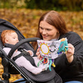 A woman smiles while showing the Things with Wings Peek a Boo Book, a soft, colorful fabric book with playful designs and attached toys, to a baby in a stroller outdoors. The baby gazes intently at the engaging book.